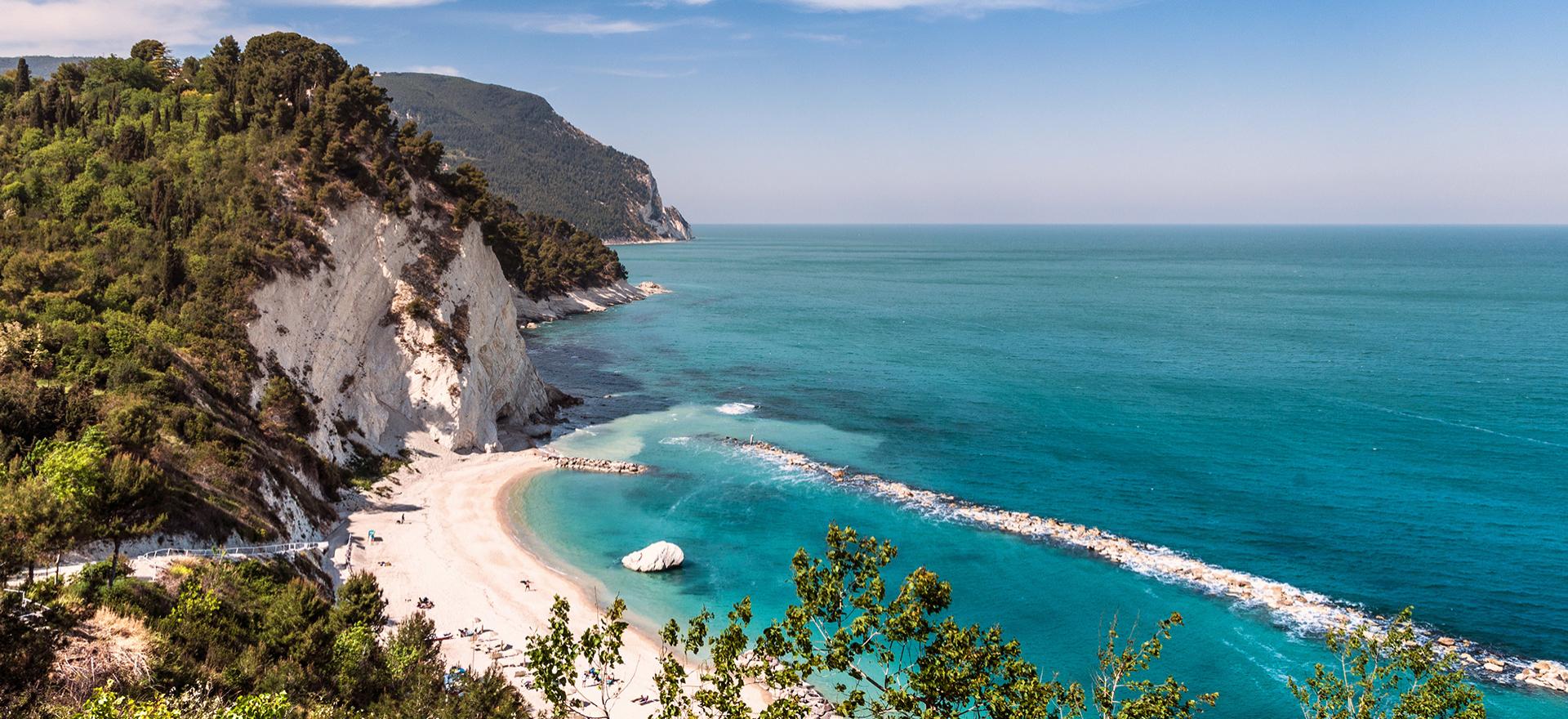 Spiaggia con sabbia bianca e mare cristallino, circondata da scogliere e vegetazione.